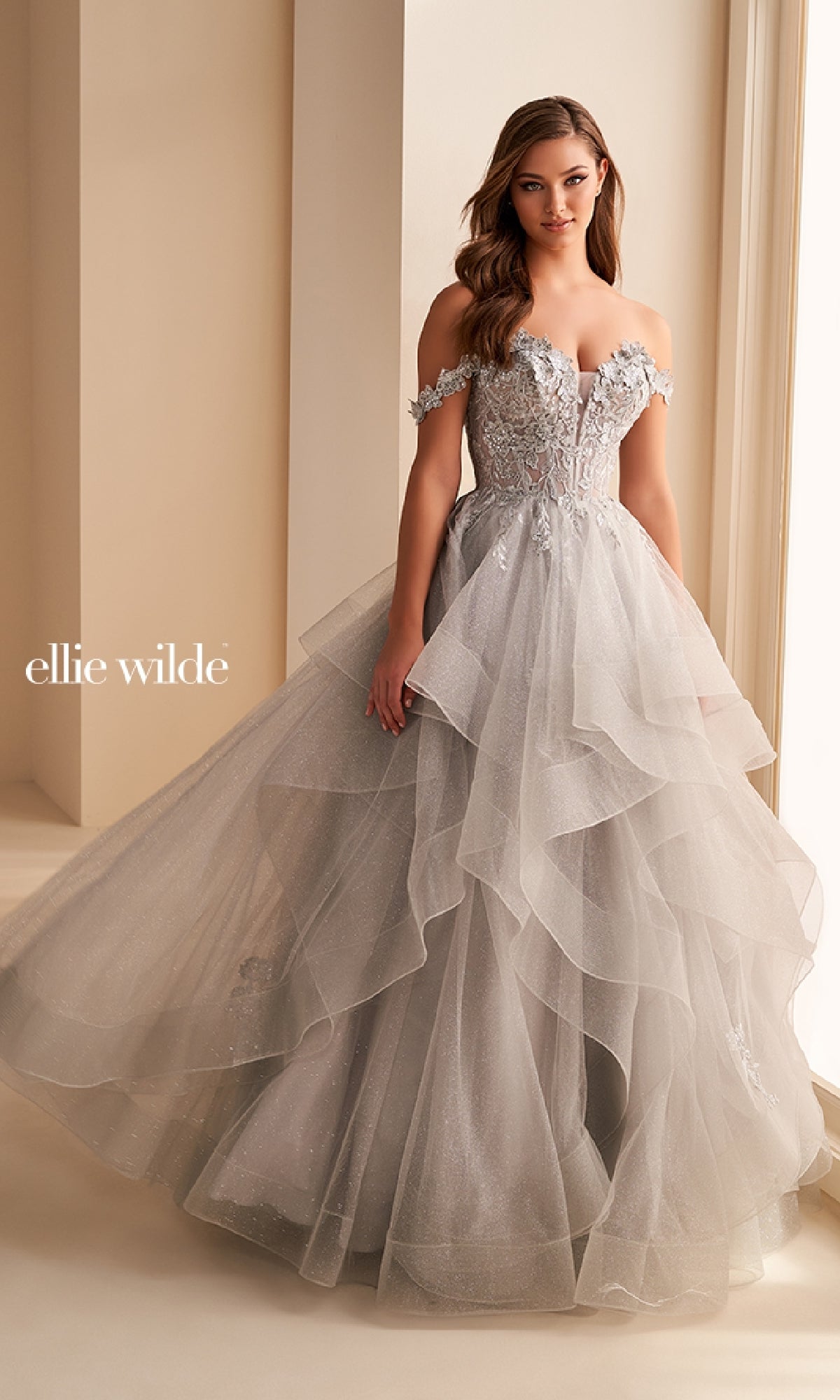 A woman stands in a softly lit room wearing the Ellie Wilde Long Designer Prom Ball Gown EW35084, an off-the-shoulder, tiered gown with floral embellishments and shimmering silver glitter tulle.